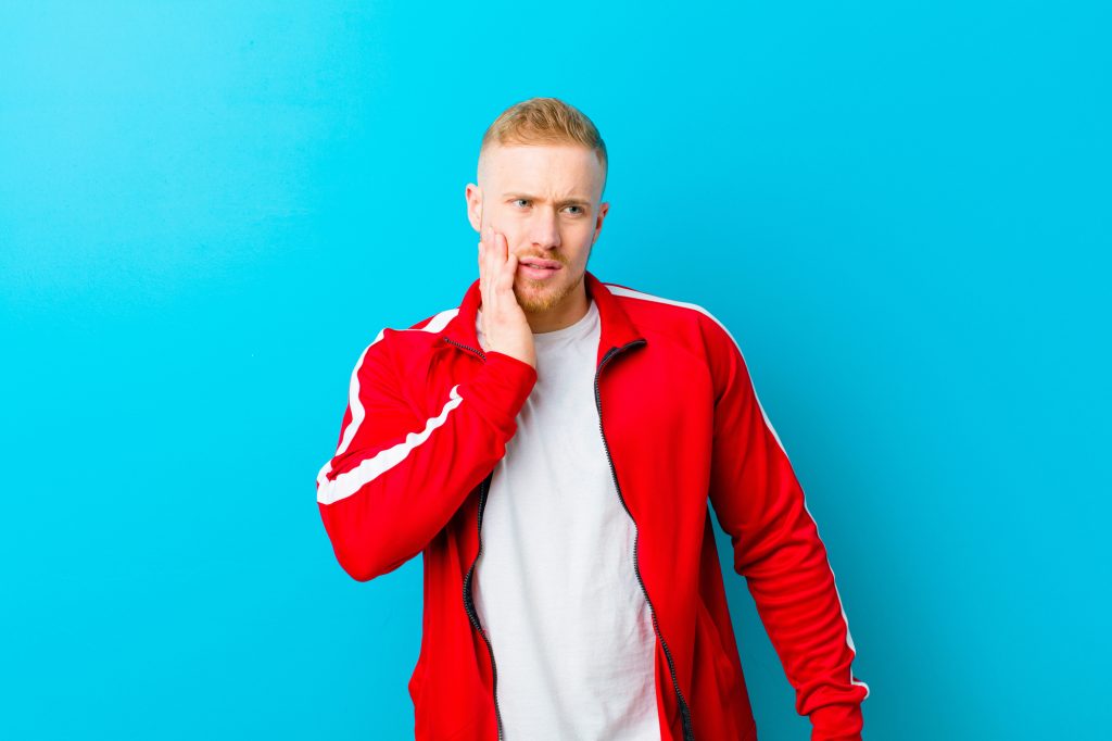 young blonde man wearing sports clothes holding cheek and suffering painful toothache, feeling ill, miserable and unhappy, looking for a dentist against blue background