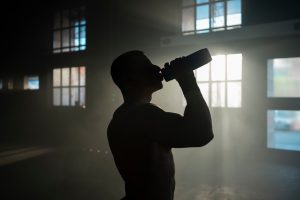 Topless muscular sportsman drinking water from a sports bottle in abandoned warehouse after doing some exercise